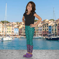 A young girl wearing kids size purple and green leopard glamour funky festival leggings stands facing forwards. She is smiling and standing in front of a red, blue and yellow circus big top tent. This design features an animal print pattern in black, grey and white, printed at a medium scale. The top 3/4 background is mint green with the bottom 1/4 purple