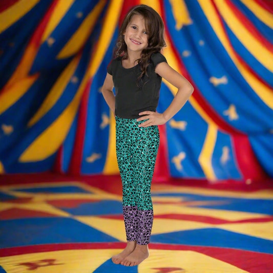 A young girl wearing kids size purple and green leopard glamour funky festival leggings stands facing forwards. She is smiling and standing in front of a red, blue and yellow circus big top tent. This design features an animal print pattern in black, grey and white, printed at a medium scale. The top 3/4 background is mint green with the bottom 1/4 purple
