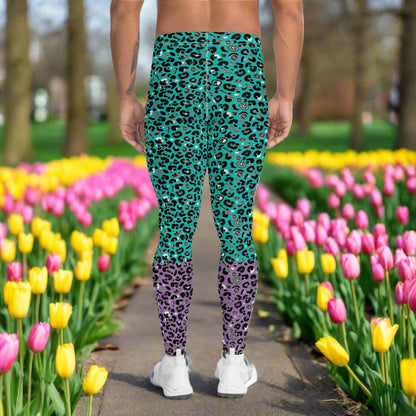 Man from the waist down wearing mens Purple and Green Leopard Glamour funky festival yoga leggings standing facing away with his hands by his sides. The man is standing in a park in spring with pink and yellow tulips flowering and trees in the background. This design features an animal print pattern in black, grey and white, printed at a medium scale. The top 3/4 background is mint green with the bottom 1/4 purple