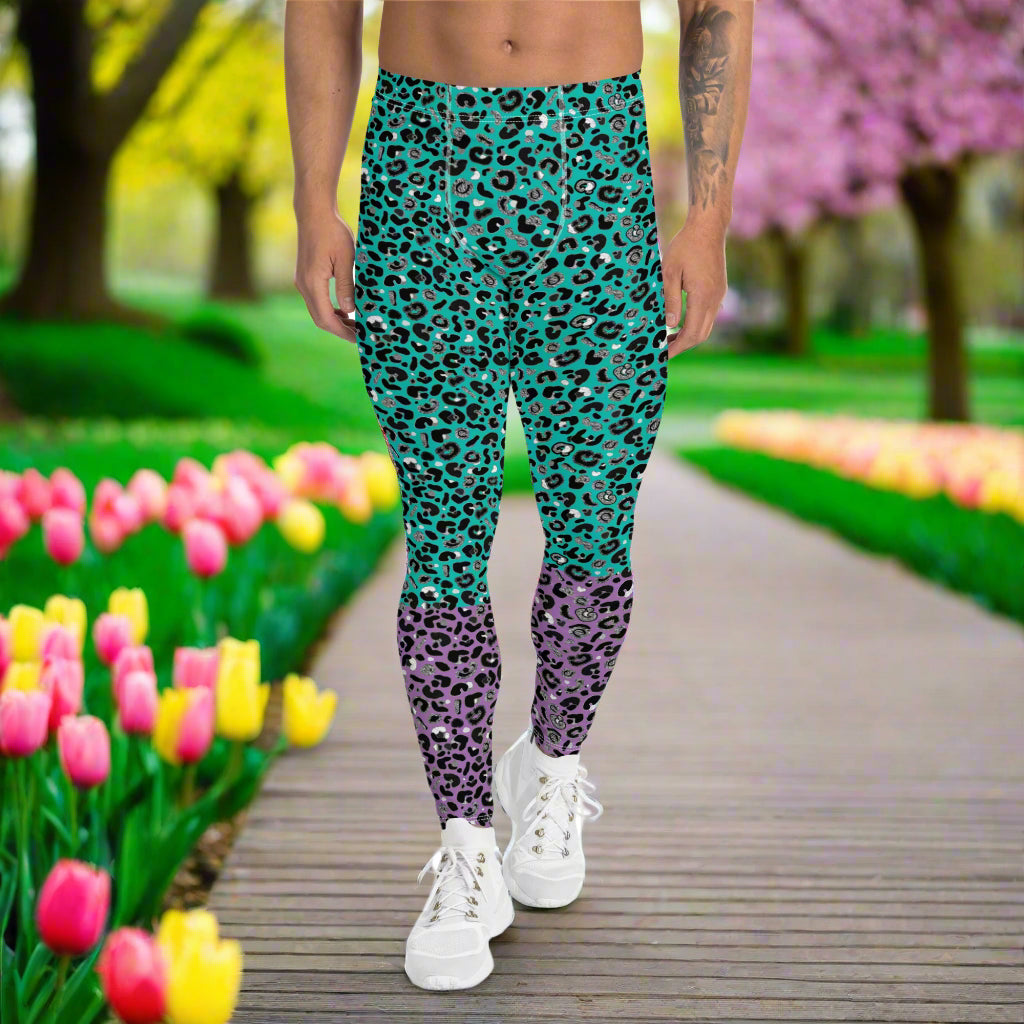 Man from the waist down wearing mens Purple and Green Leopard Glamour funky festival yoga leggings standing facing forwards with his hands by his sides. The man is standing in a park in spring with blossoming trees and pink and yellow tulips flowering. This design features an animal print pattern in black, grey and white, printed at a medium scale. The top 3/4 background is mint green with the bottom 1/4 purple