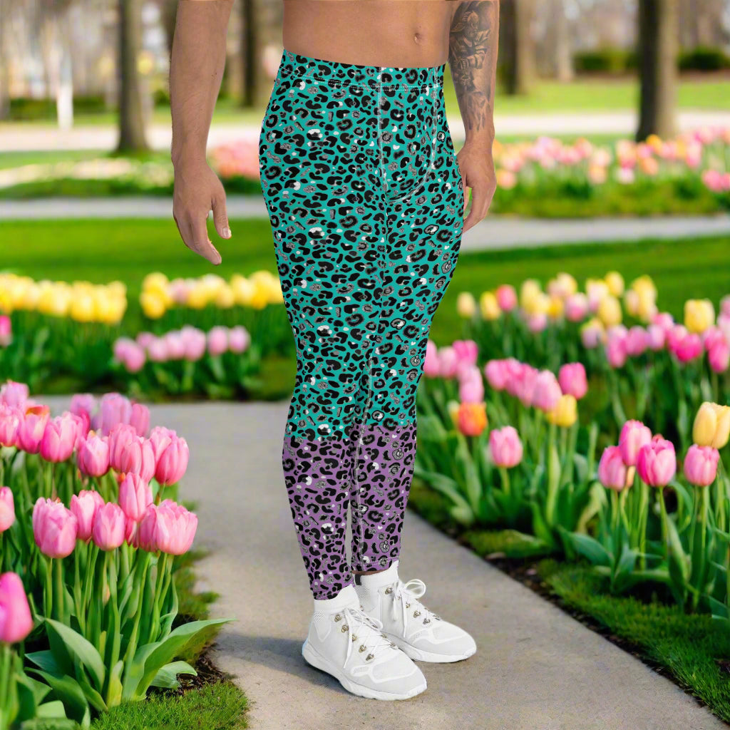 Man from the waist down wearing mens Purple and Green Leopard Glamour funky festival yoga leggings standing facing to the right with his hands by his sides. The man is standing in a park in spring with pink and yellow tulips flowering and trees in the background. This design features an animal print pattern in black, grey and white, printed at a medium scale. The top 3/4 background is mint green with the bottom 1/4 purple