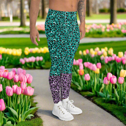 Man from the waist down wearing mens Purple and Green Leopard Glamour funky festival yoga leggings standing facing to the right with his hands by his sides. The man is standing in a park in spring with pink and yellow tulips flowering and trees in the background. This design features an animal print pattern in black, grey and white, printed at a medium scale. The top 3/4 background is mint green with the bottom 1/4 purple