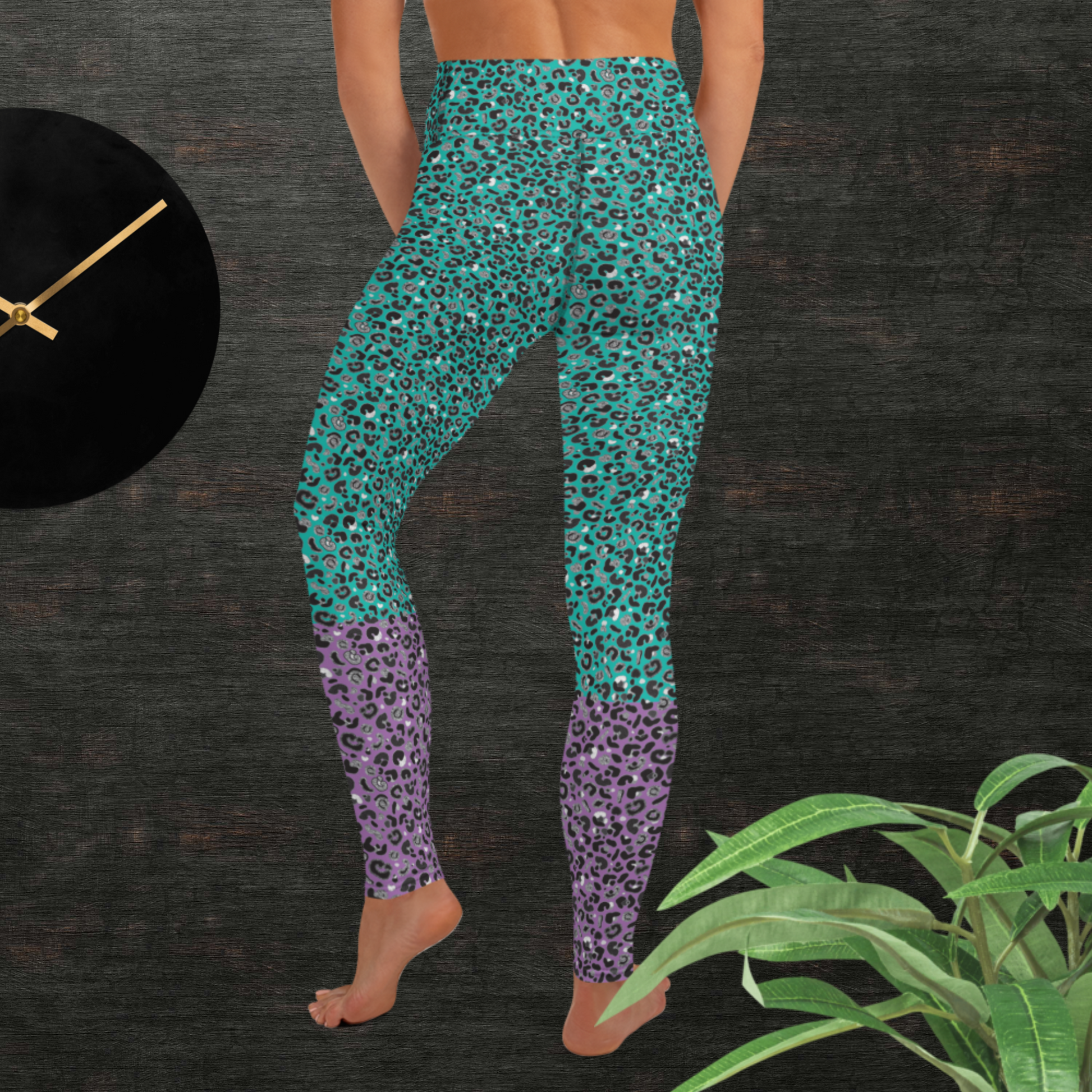 Woman from the waist down wearing high-waist Purple and Green Leopard Glamour funky festival yoga leggings standing facing backwards with her hands in front of her. The woman is standing in front of a textured grey background with a black and gold wall clock to the left and a partially visible leafy plant on the floor in the bottom right.  This design features an animal print pattern in black, grey and white, printed at a medium scale. The top 3/4 background is mint green with the bottom 1/4 purple