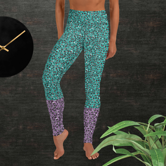 Woman from the waist down wearing high-waist Purple and Green Leopard Glamour funky festival yoga leggings standing facing forwards with her hands by her sides. The woman is standing in front of a textured grey background with a black and gold wall clock to the left and a partially visible leafy plant on the floor in the bottom right.  This design features an animal print pattern in black, grey and white, printed at a medium scale. The top 3/4 background is mint green with the bottom 1/4 purple