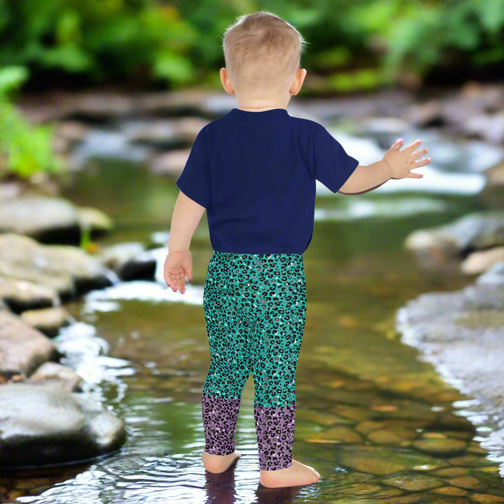 A toddler wearing kids size purple and green leopard glamour funky festival leggings and a navy t-shirt faces away. He is padding in a very shallow stream which winds from the front of the picture towards the back, surrounded by rocks and stones and greenery. This design features an animal print pattern in black, grey and white, printed at a medium scale. The top 3/4 background is mint green with the bottom 1/4 purple