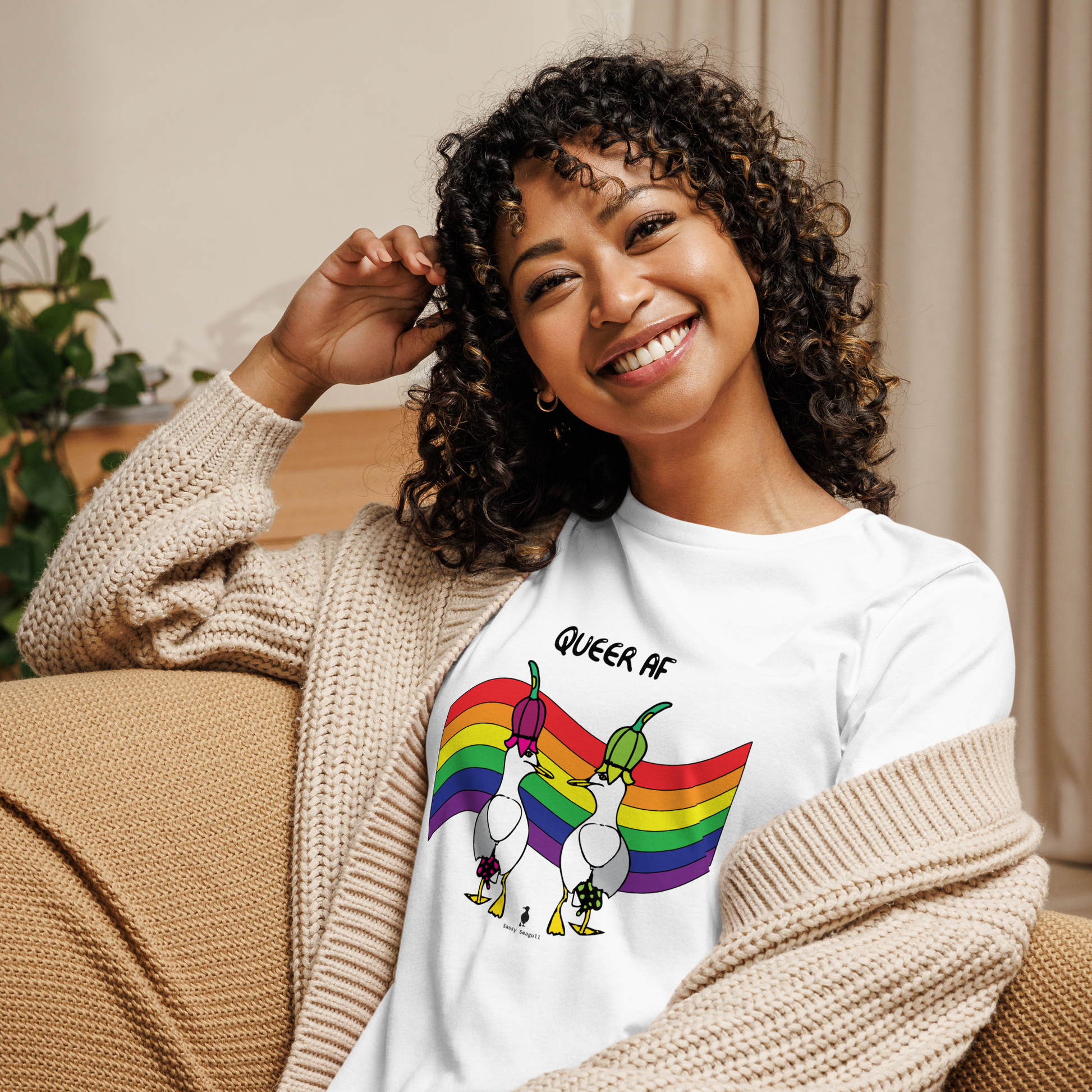 Woman sitting relaxing and smiling at the camera. She is in a living area on comfy seating, wearing a fitted white Sassy Seagull Queer AF LGBTQ+ Pride T-Shirt and a cream knitted cardigan. The t-shirt is white with short sleeves. The graphic shows two cartoon style seagulls wearing colourful hats walking away in front of an LGBTQIA pride flag, underneath the wording Queer AF. It is a bold playful design for everyday wear.