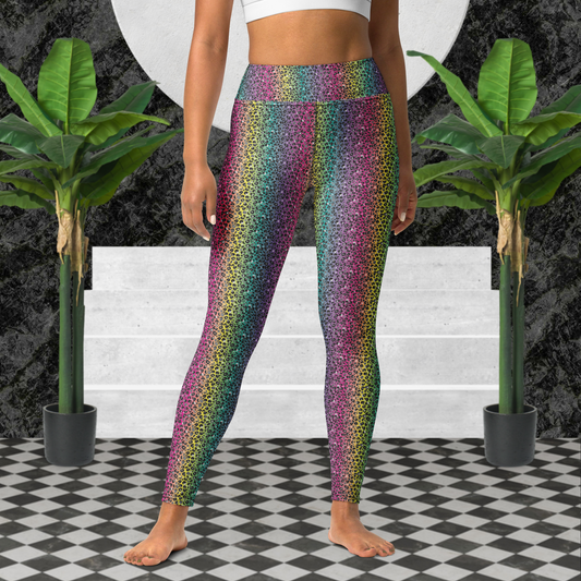 Woman from the waist down wearing high-waist Rainbow Leopard Glamour funky festival yoga leggings standing facing forwards with her hands by her sides. The woman is standing on a black and white tiled floor in front of some white steps and a black and grey marble background. There is a leafy plant on the floor at each side of the steps. The rainbow leopard glamour design is an animal print pattern in black, grey and white, printed at a small scale against a blended rainbow background