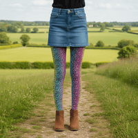 A young person pictured from the waist down wearing rainbow leopard glamour leggings and white trainers. They are facing forwards, standing on a wooden floor in a rustic cafe environment with plants and fairy lights hanging from the ceiling. The rainbow leopard glamour design is an animal print pattern in black, grey and white, printed at a small scale against a blended rainbow background
