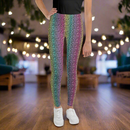 A young person pictured from the waist down wearing rainbow leopard glamour leggings and white trainers. They are facing forwards, standing on a wooden floor in a rustic cafe environment with plants and fairy lights hanging from the ceiling. The rainbow leopard glamour design is an animal print pattern in black, grey and white, printed at a small scale against a blended rainbow background