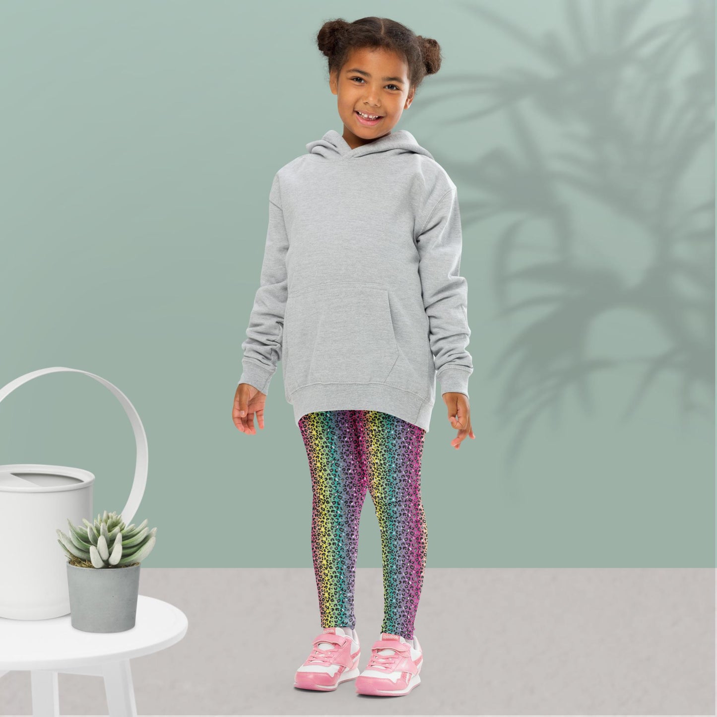 A young girl wearing kids size rainbow leopard glamour funky festival leggings and an oversized grey hoodie, stands facing forwards. She is smiling and standing in front of a pastel duck egg wall with a shadow of a large spidery plant behind her. In the front left is a small white table with a small plant and white watering can on top. The rainbow leopard glamour design is an animal print pattern in black, grey and white, printed at a small scale against a blended rainbow background