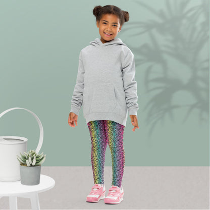 A young girl wearing kids size rainbow leopard glamour funky festival leggings and an oversized grey hoodie, stands facing forwards. She is smiling and standing in front of a pastel duck egg wall with a shadow of a large spidery plant behind her. In the front left is a small white table with a small plant and white watering can on top. The rainbow leopard glamour design is an animal print pattern in black, grey and white, printed at a small scale against a blended rainbow background