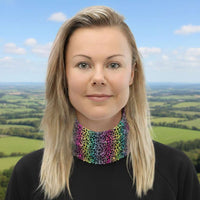 Woman with long blonde hair facing to the front wearing a grey long sleeve top and a vibrant rainbow leopard glamour neck gaiter around her neck. In the background are some cafe tables and plants. The rainbow leopard glamour design is an animal print pattern in black, grey and white, printed at a small scale against a blended rainbow background