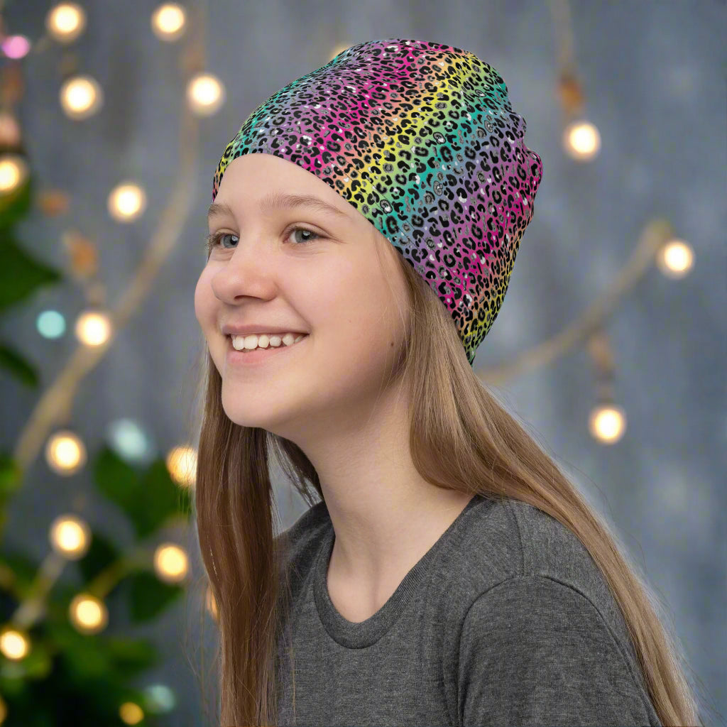 Girl with long dark blonde hair facing to the left wearing a vibrant rainbow leopard glamour neck gaiter as a beanie hat. In the background are some fairy lights and plants. The rainbow leopard glamour design is an animal print pattern in black, grey and white, printed at a small scale against a blended rainbow background
