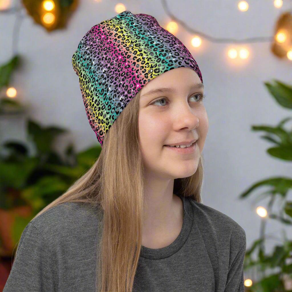 Girl with long dark blonde hair facing to the right wearing a vibrant rainbow leopard glamour neck gaiter as a beanie hat. In the background are some fairy lights and plants. The rainbow leopard glamour design is an animal print pattern in black, grey and white, printed at a small scale against a blended rainbow background