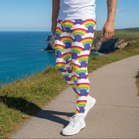 Man from the waist down wearing men's Rainbow Love funky festival yoga leggings standing facing to the left with his hands by his sides. The man is standing on a dance floor with colourful stage light spots on the floor. The background is hazy mixed with glow from the coloured lights. The rainbow love pattern features repeating rainbows and white fluffy clouds with a small pink heart beneath each rainbow. The background colour is royal sky blue