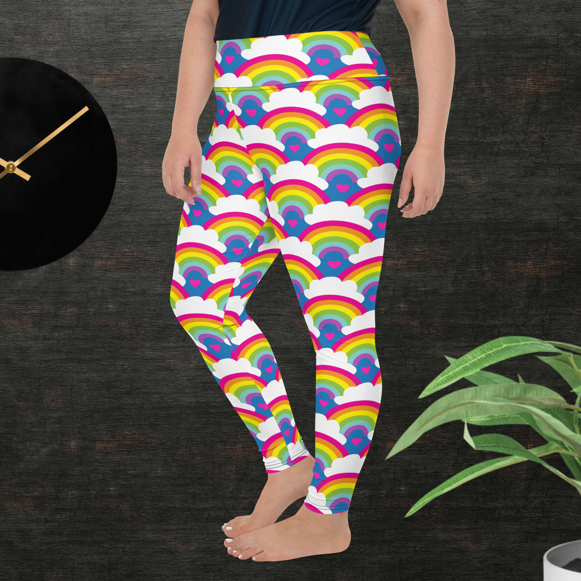 A curvy woman from the waist down wearing plus-size high-waist Rainbow Love funky festival yoga leggings standing facing to the left with her hands by her sides. The woman is standing in front of a textured grey background with a black and gold wall clock to the left and a partially visible leafy plant on the floor in the bottom right. The rainbow love pattern features repeating rainbows and white fluffy clouds with a small pink heart beneath each rainbow. The background colour is royal sky blue