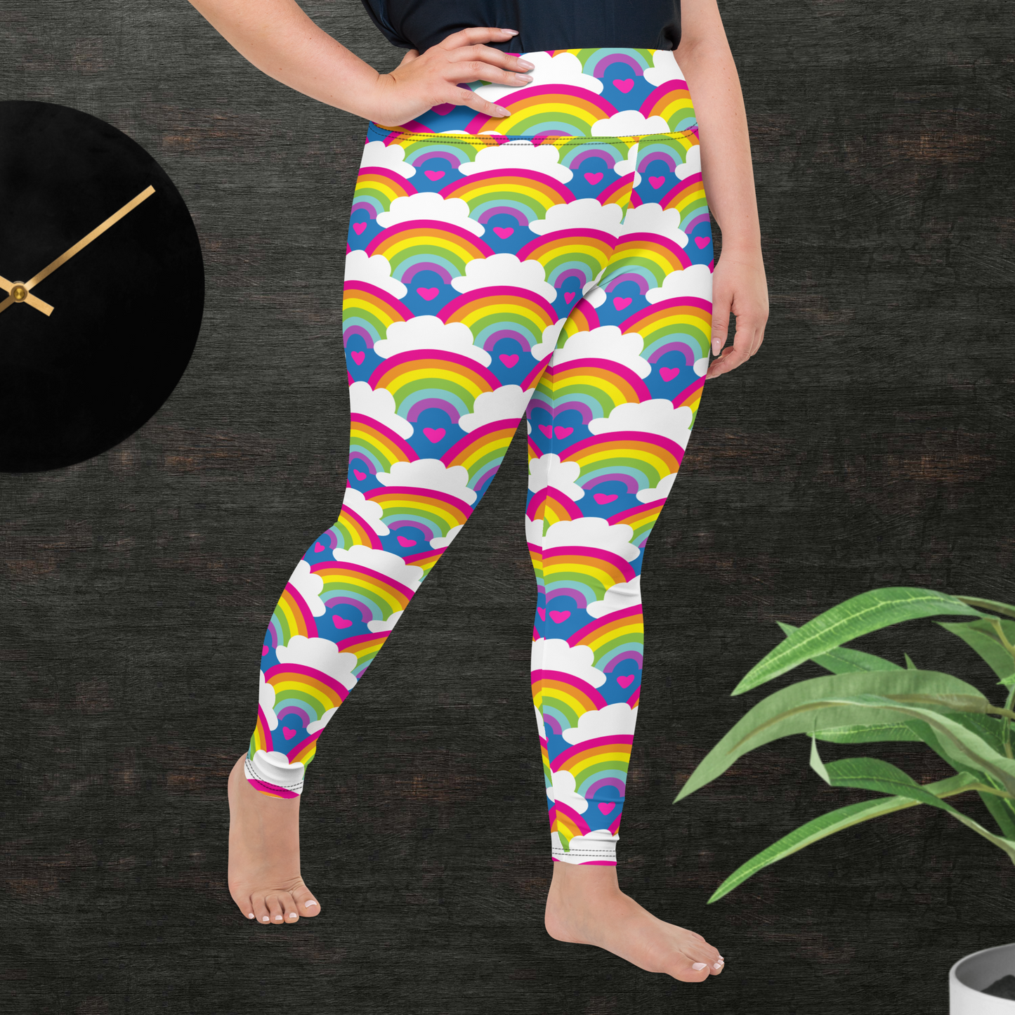 A curvy woman from the waist down wearing plus-size high-waist Rainbow Love funky festival yoga leggings standing facing to the right with her right hand on her hip. The woman is standing in front of a textured grey background with a black and gold wall clock to the left and a partially visible leafy plant on the floor in the bottom right. The rainbow love pattern features repeating rainbows and white fluffy clouds with a small pink heart beneath each rainbow. The background colour is royal sky blue