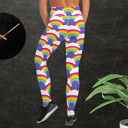 Woman from the waist down wearing high-waist Rainbow Love funky festival yoga leggings standing facing backwards with her hands in front of her. The woman is standing in front of a textured grey background with a black and gold wall clock to the left and a partially visible leafy plant on the floor in the bottom right. The rainbow love pattern features repeating rainbows and white fluffy clouds with a small pink heart beneath each rainbow. The background colour is royal sky blue