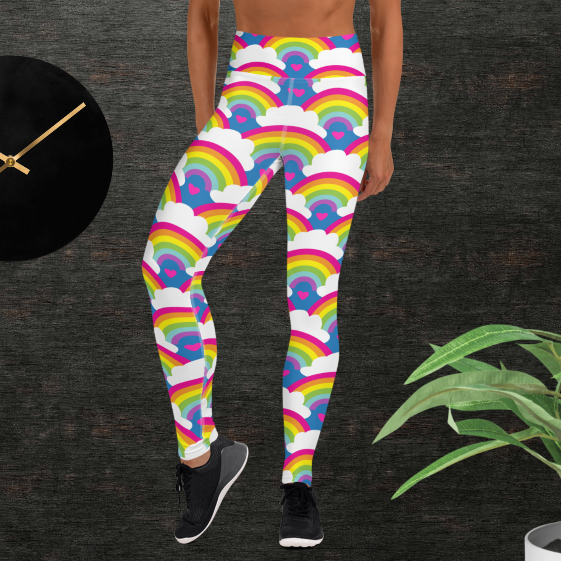 Woman from the waist down wearing high-waist Rainbow Love funky festival yoga leggings standing facing forwards with her hands by her sides. The woman is standing in front of a textured grey background with a black and gold wall clock to the left and a partially visible leafy plant on the floor in the bottom right. The rainbow love pattern features repeating rainbows and white fluffy clouds with a small pink heart beneath each rainbow. The background colour is royal sky blue