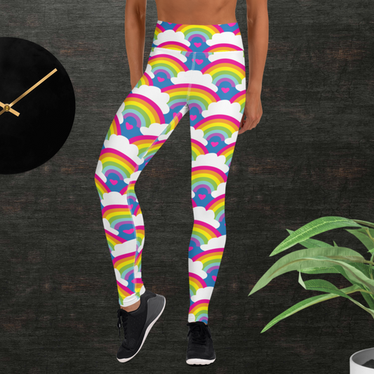 Woman from the waist down wearing high-waist Rainbow Love funky festival yoga leggings standing facing forwards with her hands by her sides. The woman is standing in front of a textured grey background with a black and gold wall clock to the left and a partially visible leafy plant on the floor in the bottom right. The rainbow love pattern features repeating rainbows and white fluffy clouds with a small pink heart beneath each rainbow. The background colour is royal sky blue
