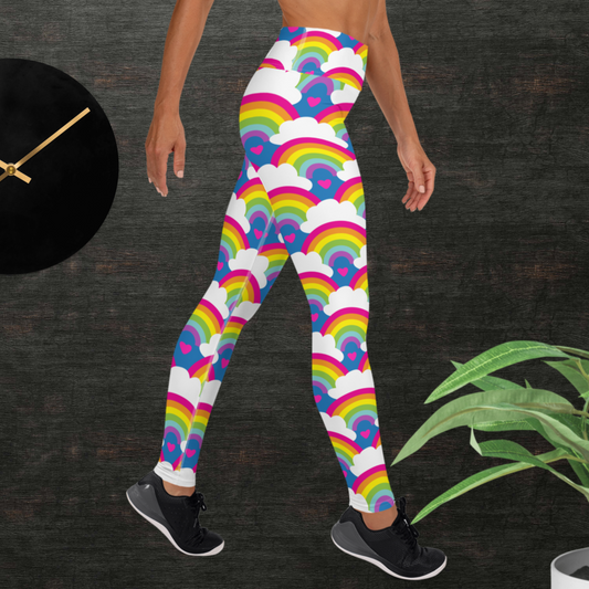 Woman from the waist down wearing high-waist Rainbow Love funky festival yoga leggings standing facing to the right with her hands by her sides. The woman is standing in front of a textured grey background with a black and gold wall clock to the left and a partially visible leafy plant on the floor in the bottom right. The rainbow love pattern features repeating rainbows and white fluffy clouds with a small pink heart beneath each rainbow. The background colour is royal sky blue