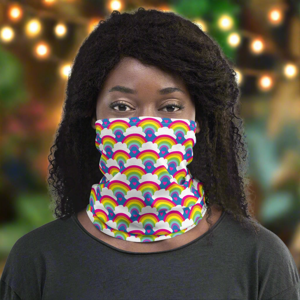 Woman with afro hair facing to the front wearing a grey top and a vibrant rainbow love neck gaiter around her neck, covering her mouth and nose. In the background are some fairy lights and plants. The rainbow love pattern features repeating rainbows and white fluffy clouds with a small pink heart beneath each rainbow. The background colour is royal sky blue.