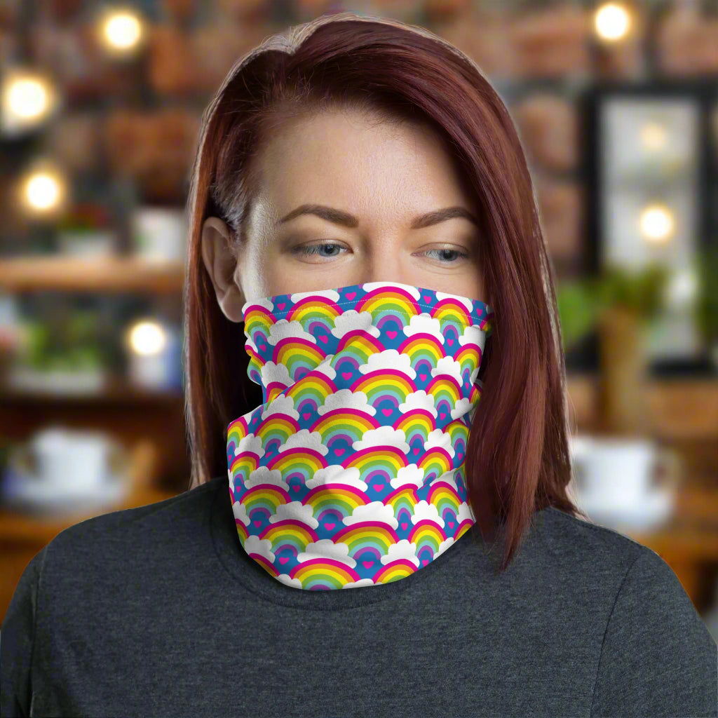 Woman with red hair looking to the right wearing a grey top and a vibrant rainbow love neck gaiter around her neck, covering her mouth and nose. In the background are some fairy lights and plants. The rainbow love pattern features repeating rainbows and white fluffy clouds with a small pink heart beneath each rainbow. The background colour is royal sky blue.