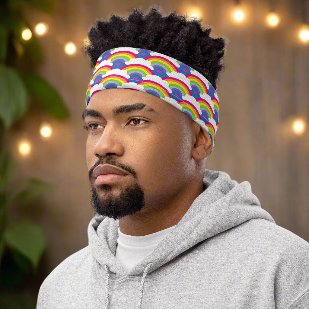Man with short afro hair facing to the left wearing a grey top and a vibrant rainbow love neck gaiter around his head as a headband. In the background are some fairy lights and plants. The rainbow love pattern features repeating rainbows and white fluffy clouds with a small pink heart beneath each rainbow. The background colour is royal sky blue.