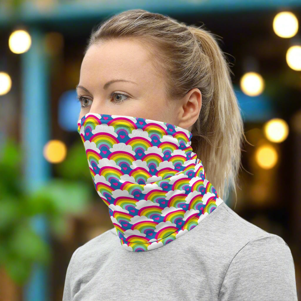 Woman with blonde hair facing to the left wearing a grey top and a vibrant rainbow love neck gaiter around her neck, covering her mouth and nose. In the background are some fairy lights and plants. The rainbow love pattern features repeating rainbows and white fluffy clouds with a small pink heart beneath each rainbow. The background colour is royal sky blue.
