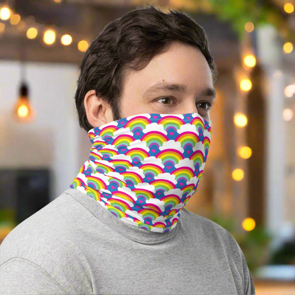 Man with dark hair facing to the right wearing a grey top and a vibrant rainbow love neck gaiter around his neck, covering his mouth and nose. In the background are some fairy lights and plants. The rainbow love pattern features repeating rainbows and white fluffy clouds with a small pink heart beneath each rainbow. The background colour is royal sky blue.