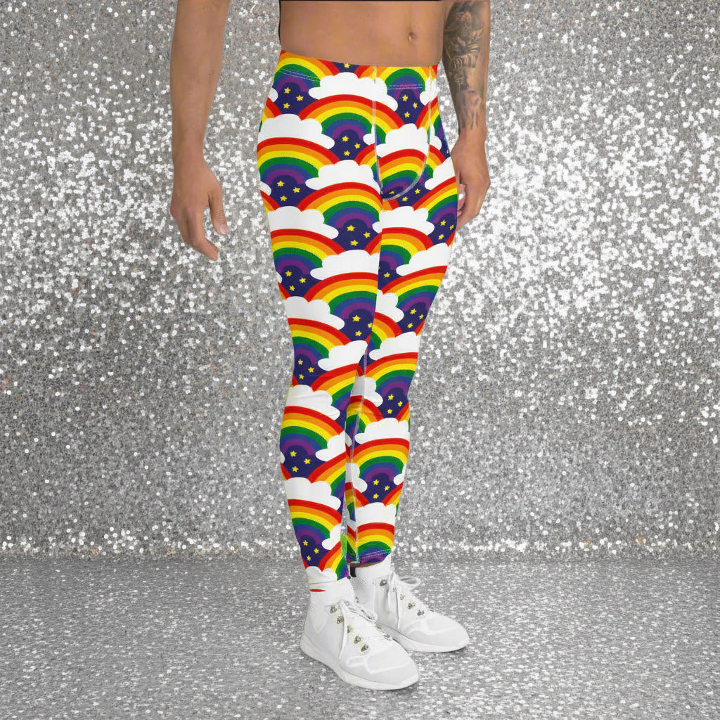 Man from the waist down wearing men's Rainbow Sparkle funky festival yoga leggings standing facing  to the right with his hands by his sides. The man is standing in a nightclub with colourful pink and purple lighting in the background. The rainbow sparkle pattern features repeating rainbows and white fluffy clouds with three small yellow stars beneath each rainbow. The background colour is navy blue