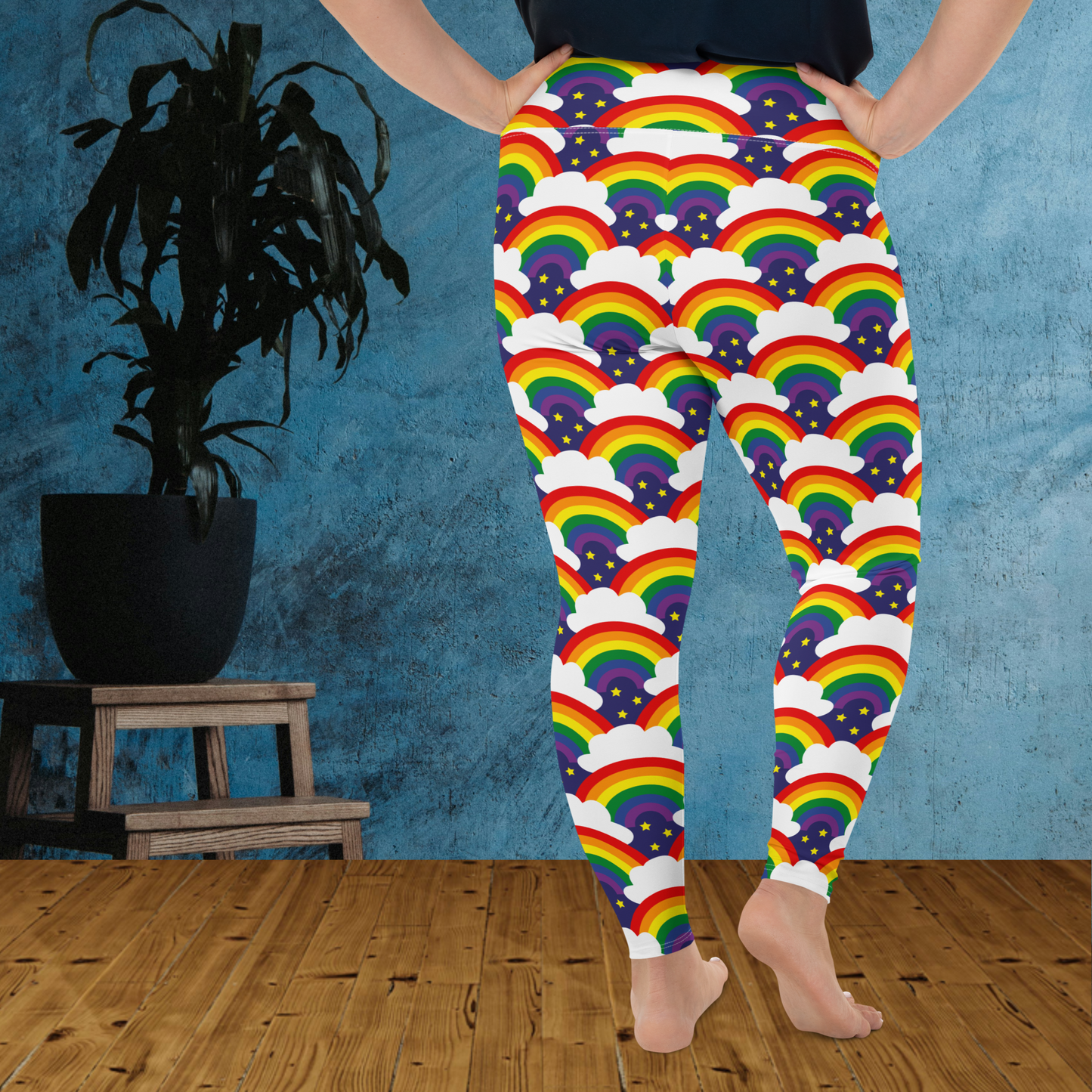 A curvy woman from the waist down wearing plus-size high-waist Rainbow Sparkle Pride funky festival yoga leggings standing facing backwards with her hands on her hips. The woman is standing on a wooden floor in front of a textured blue background with a leafy plant in a black pot on a small plant stand to the left of the image. The rainbow sparkle pattern features repeating rainbows and white fluffy clouds with three small yellow stars beneath each rainbow. The background colour is navy blue