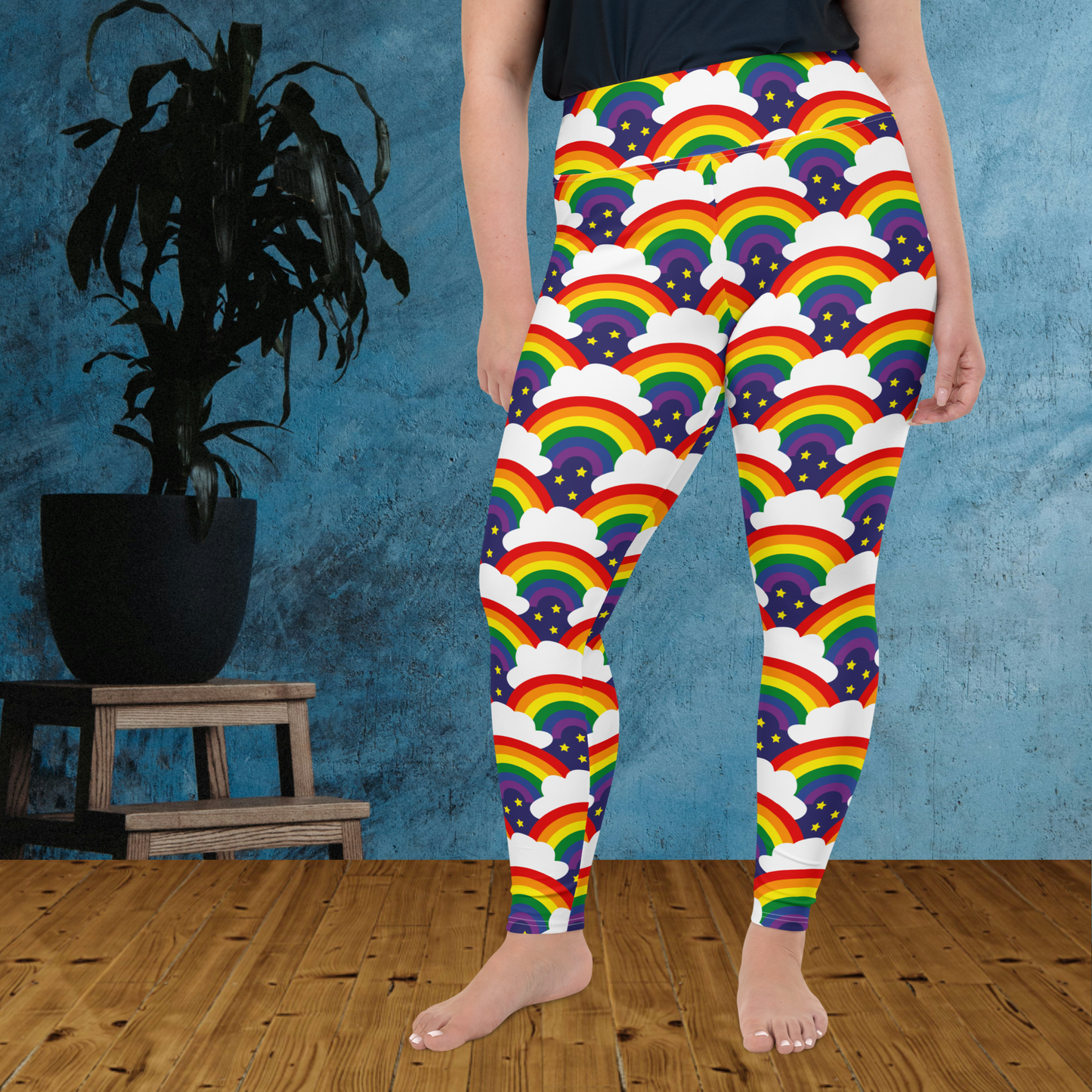 A curvy woman from the waist down wearing plus-size high-waist Rainbow Sparkle Pride funky festival yoga leggings standing facing forwards with her hands by her sides. The woman is standing on a wooden floor in front of a textured blue background with a leafy plant in a black pot on a small plant stand to the left of the image. The rainbow sparkle pattern features repeating rainbows and white fluffy clouds with three small yellow stars beneath each rainbow. The background colour is navy blue