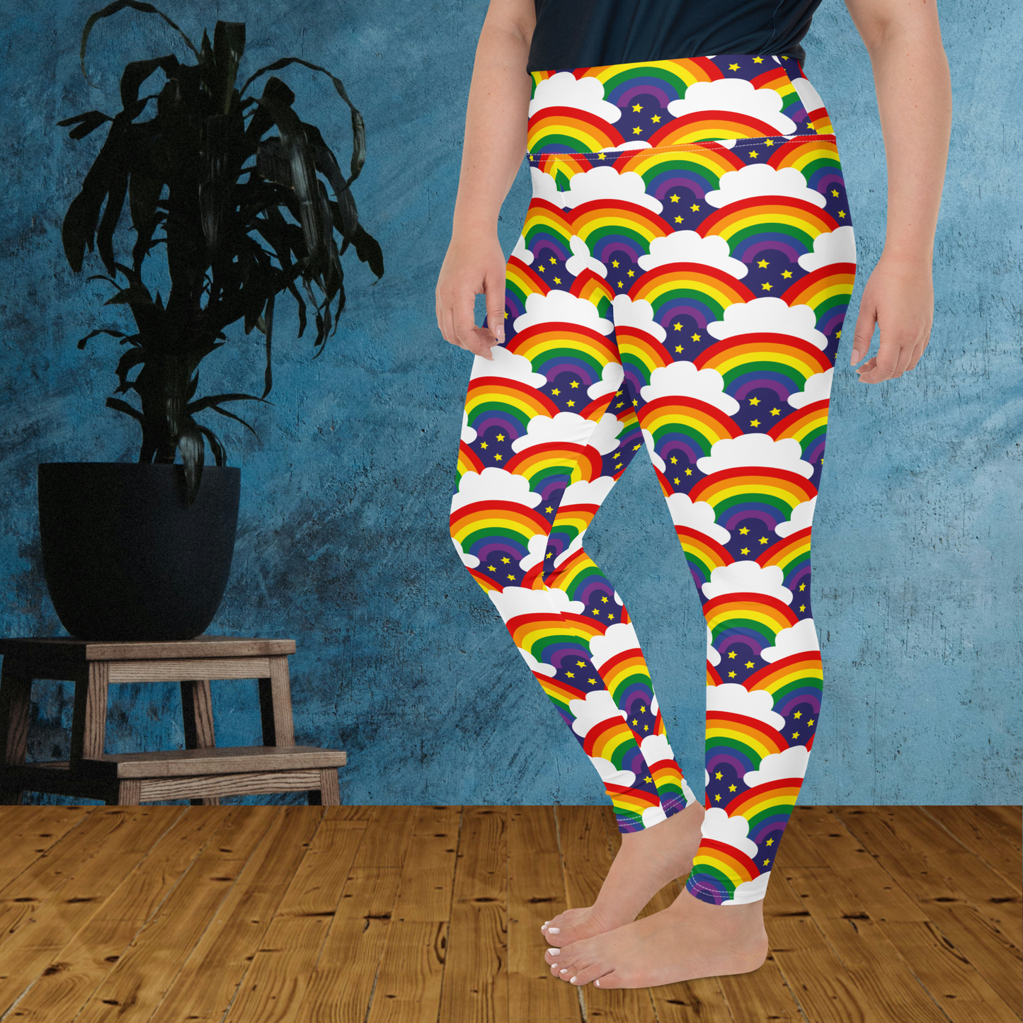 A curvy woman from the waist down wearing plus-size high-waist Rainbow Sparkle Pride funky festival yoga leggings standing facing to the left with her hands by her sides. The woman is standing on a wooden floor in front of a textured blue background with a leafy plant in a black pot on a small plant stand to the left of the image. The rainbow sparkle pattern features repeating rainbows and white fluffy clouds with three small yellow stars beneath each rainbow. The background colour is navy blue