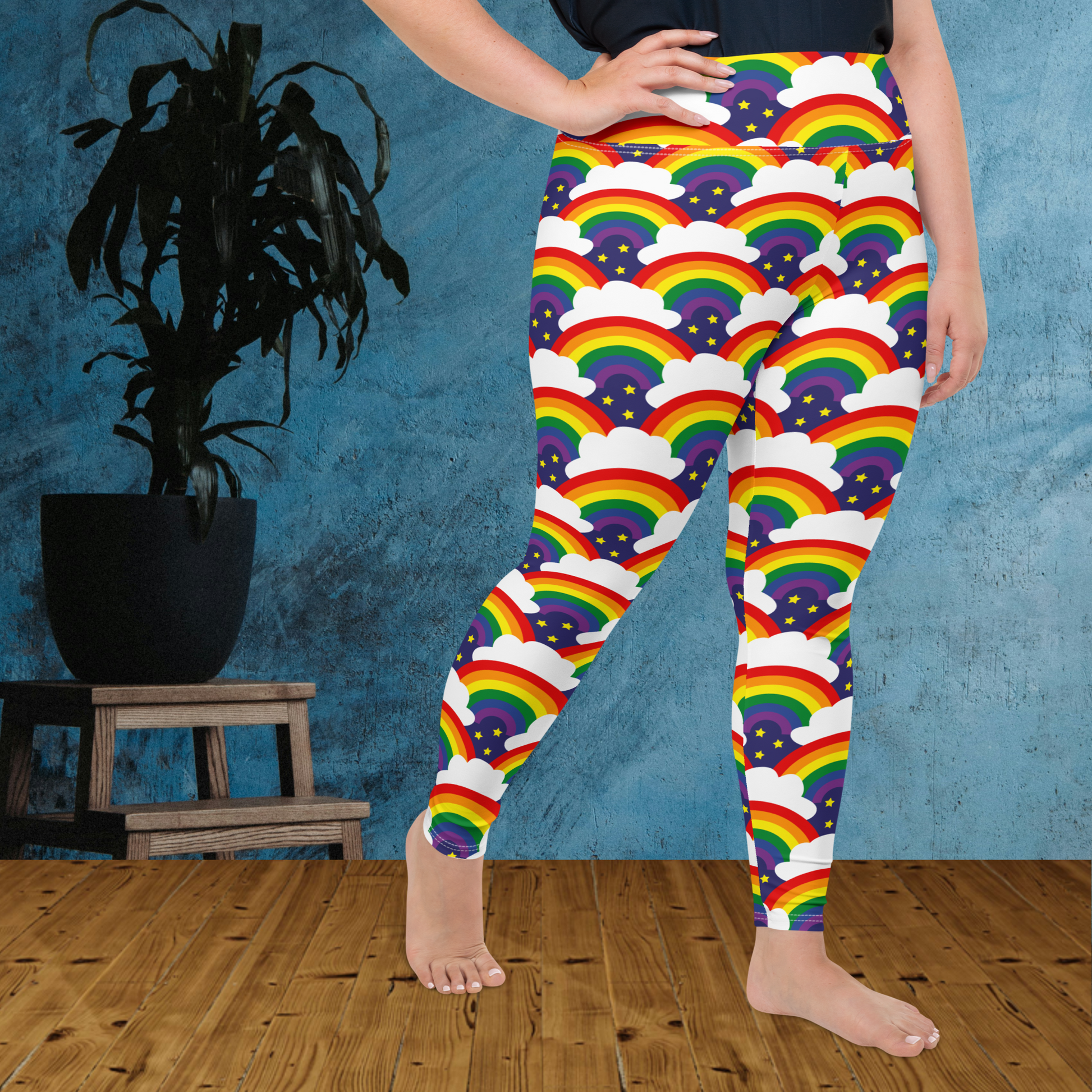 A curvy woman from the waist down wearing plus-size high-waist Rainbow Sparkle Pride funky festival yoga leggings standing facing to the right with her right hand on her hip. The woman is standing on a wooden floor in front of a textured blue background with a leafy plant in a black pot on a small plant stand to the left of the image. The rainbow sparkle pattern features repeating rainbows and white fluffy clouds with three small yellow stars beneath each rainbow. The background colour is navy blue