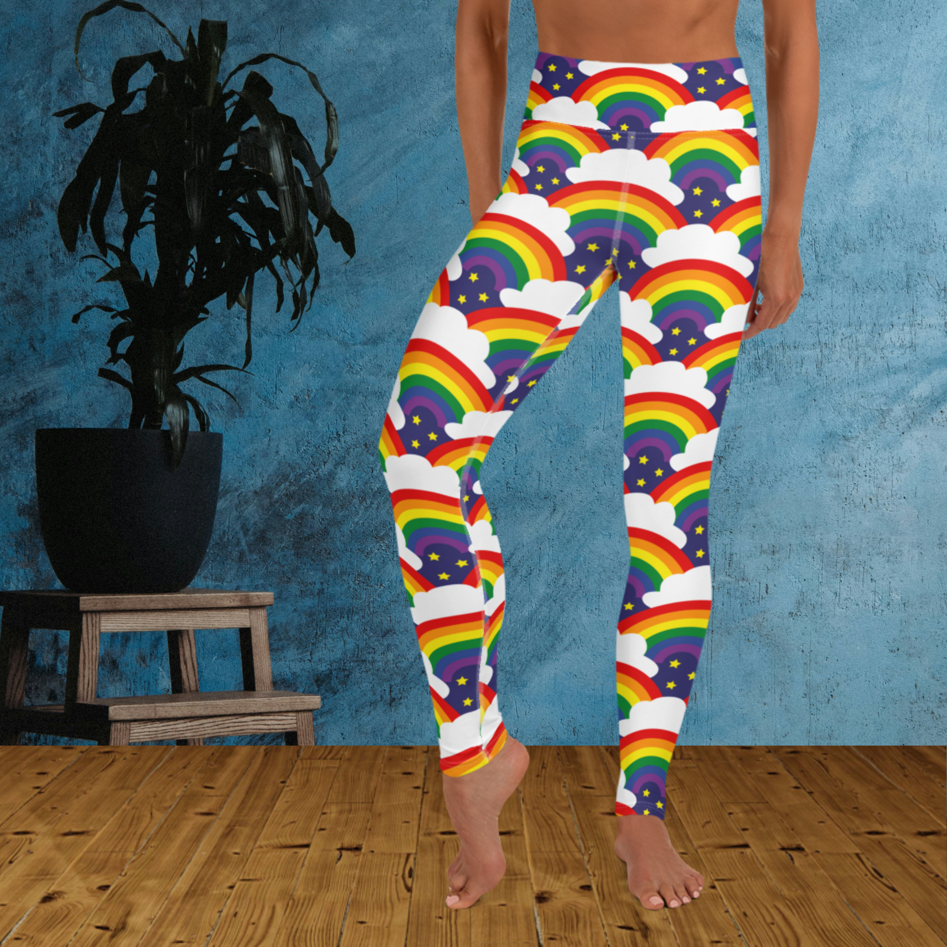 Woman from the waist down wearing high-waist Rainbow Sparkle Pride funky festival yoga leggings standing facing forwards with her hands by her sides. The woman is standing on a wooden floor in front of a textured blue background with a leafy plant in a black pot on a small plant stand to the left of the image. The rainbow sparkle pattern features repeating rainbows and white fluffy clouds with three small yellow stars beneath each rainbow. The background colour is navy blue