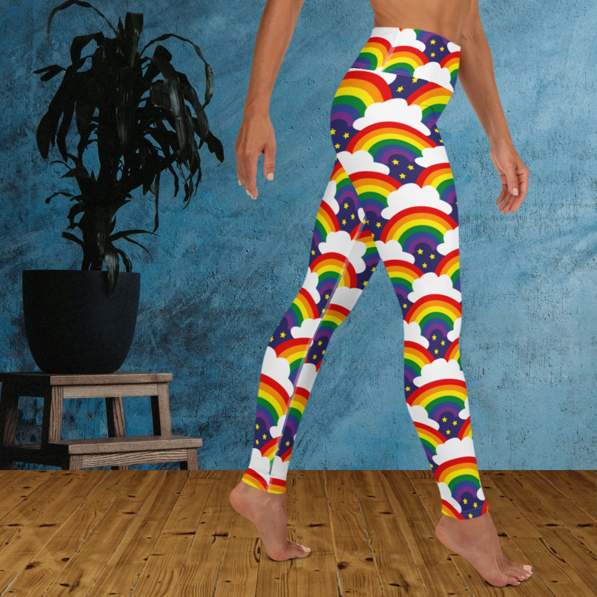 Woman from the waist down wearing high-waist Rainbow Sparkle Pride funky festival yoga leggings standing facing to the right with her hands by her sides. The woman is standing on a wooden floor in front of a textured blue background with a leafy plant in a black pot on a small plant stand to the left of the image. The rainbow sparkle pattern features repeating rainbows and white fluffy clouds with three small yellow stars beneath each rainbow. The background colour is navy blue