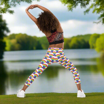 A young girl wearing kids size royal blue rainbow love funky festival leggings, poses facing away. She is posing in front of a calm lake, surrounded by green trees.  The rainbow love pattern features repeating rainbows and white fluffy clouds with a small pink heart beneath each rainbow. The background colour is royal sky blue.