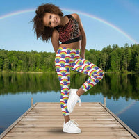 A young girl wearing kids size royal blue rainbow love funky festival leggings, poses on one leg facing forwards. She is smiling and standing in front of a calm lake, surrounded by green trees which reflect across the lake.  The rainbow love pattern features repeating rainbows and white fluffy clouds with a small pink heart beneath each rainbow. The background colour is royal sky blue.
