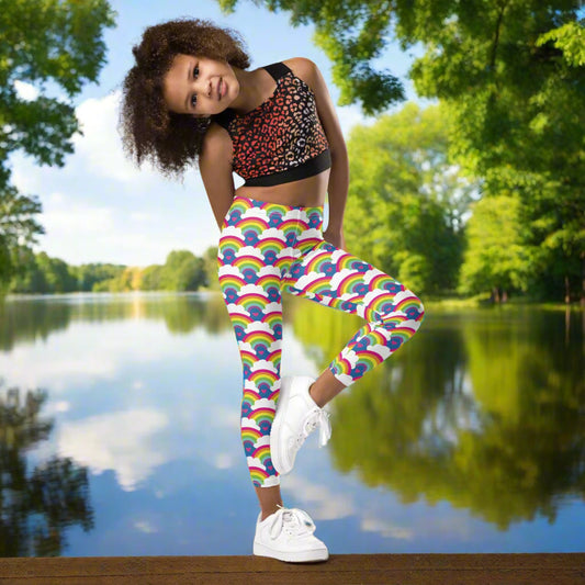 A young girl wearing kids size royal blue rainbow love funky festival leggings, poses on one leg facing forwards. She is smiling and standing in front of a calm lake, surrounded by green trees which reflect across the lake.  The rainbow love pattern features repeating rainbows and white fluffy clouds with a small pink heart beneath each rainbow. The background colour is royal sky blue.