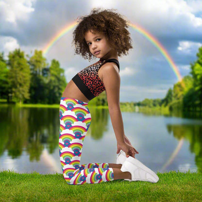 A young girl wearing kids size royal blue rainbow love funky festival leggings, poses facing to the left. She is posing in front of a calm lake, surrounded by green trees which reflect across the lake. A rainbow shines overhead also reflecting in the lake. The rainbow love pattern features repeating rainbows and white fluffy clouds with a small pink heart beneath each rainbow. The background colour is royal sky blue.