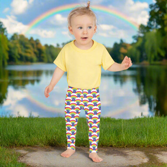 A toddler wearing kids size royal blue rainbow love funky festival leggings and a pale yellow t-shirt, stands facing forwards in front of a calm lake, surrounded by green trees which reflect across the lake. A rainbow arcs above her in the sky.  The rainbow love pattern features repeating rainbows and white fluffy clouds with a small pink heart beneath each rainbow. The background colour is royal sky blue.