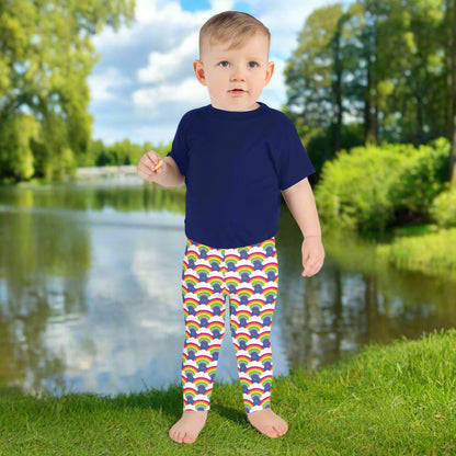 A toddler wearing kids size royal blue rainbow love funky festival leggings, and a navy t-shirt faces forwards. He is  standing in front of a calm lake, surrounded by green trees which reflect across the lake.  The rainbow love pattern features repeating rainbows and white fluffy clouds with a small pink heart beneath each rainbow. The background colour is royal sky blue.