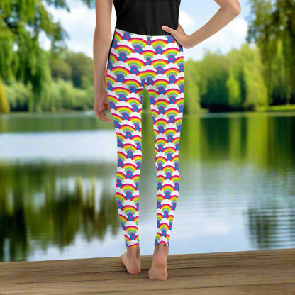 A young person from the waist down wearing youth size royal blue rainbow love funky festival leggings, stands facing away. They are standing in front of a calm lake, surrounded by green trees which reflect across the lake.  The rainbow love pattern features repeating rainbows and white fluffy clouds with a small pink heart beneath each rainbow. The background colour is royal sky blue.