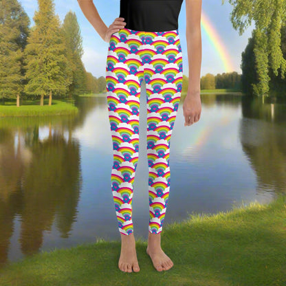 A young person from the waist down wearing youth size royal blue rainbow love funky festival leggings, stands facing forwards. They are standing in front of a calm lake, surrounded by green trees which reflect across the lake. There is a rainbow in the sky. The rainbow love pattern features repeating rainbows and white fluffy clouds with a small pink heart beneath each rainbow. The background colour is royal sky blue.