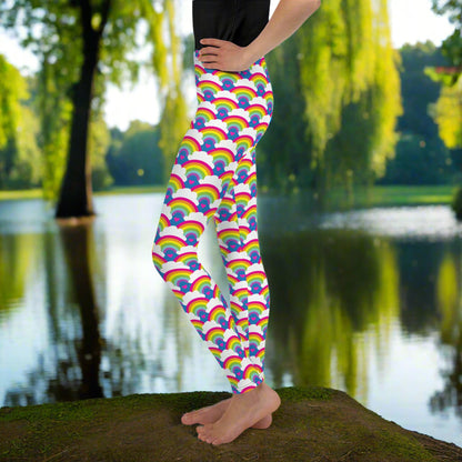 A young person from the waist down wearing youth size royal blue rainbow love funky festival leggings, stands facing to the left. They are standing in front of a calm lake, surrounded by green trees which reflect across the lake.  The rainbow love pattern features repeating rainbows and white fluffy clouds with a small pink heart beneath each rainbow. The background colour is royal sky blue.