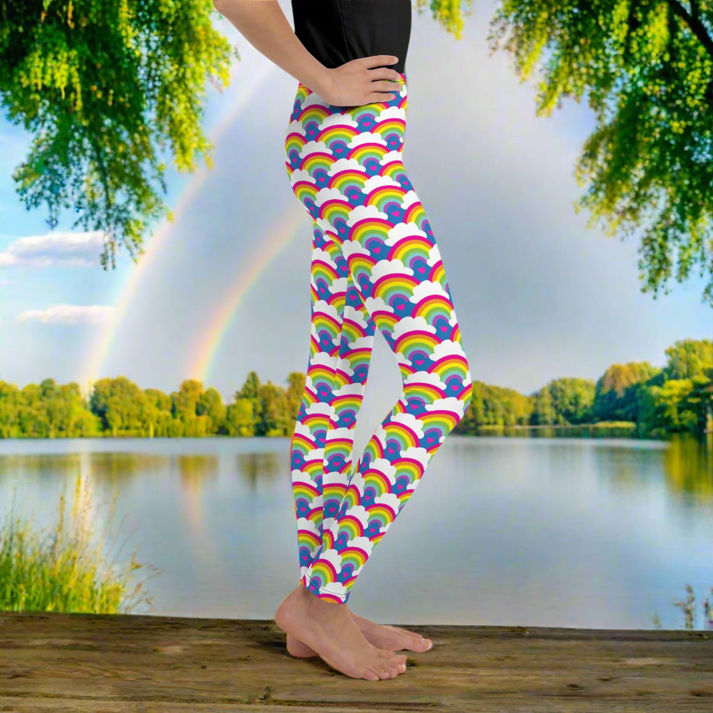 A young person from the waist down wearing youth size royal blue rainbow love funky festival leggings, stands facing to the right. They are standing in front of a calm lake, surrounded by green trees which reflect across the lake.  The rainbow love pattern features repeating rainbows and white fluffy clouds with a small pink heart beneath each rainbow. The background colour is royal sky blue.