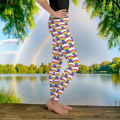 A young person from the waist down wearing youth size royal blue rainbow love funky festival leggings, stands facing to the right. They are standing in front of a calm lake, surrounded by green trees which reflect across the lake.  The rainbow love pattern features repeating rainbows and white fluffy clouds with a small pink heart beneath each rainbow. The background colour is royal sky blue.