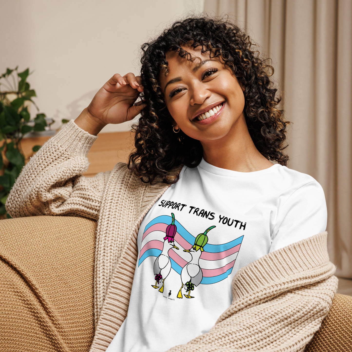 Woman sitting relaxing and smiling at the camera. She is in a living area on comfy seating, wearing a fitted white Sassy Seagull Support Trans Youth LGBTQ+ Pride T-Shirt and a cream knitted cardigan. The t-shirt is white with short sleeves. The graphic shows two cartoon style seagulls wearing colourful hats walking away in front of a trans pride flag, underneath the wording Support Trans Youth. It is a soft playful design for everyday wear.