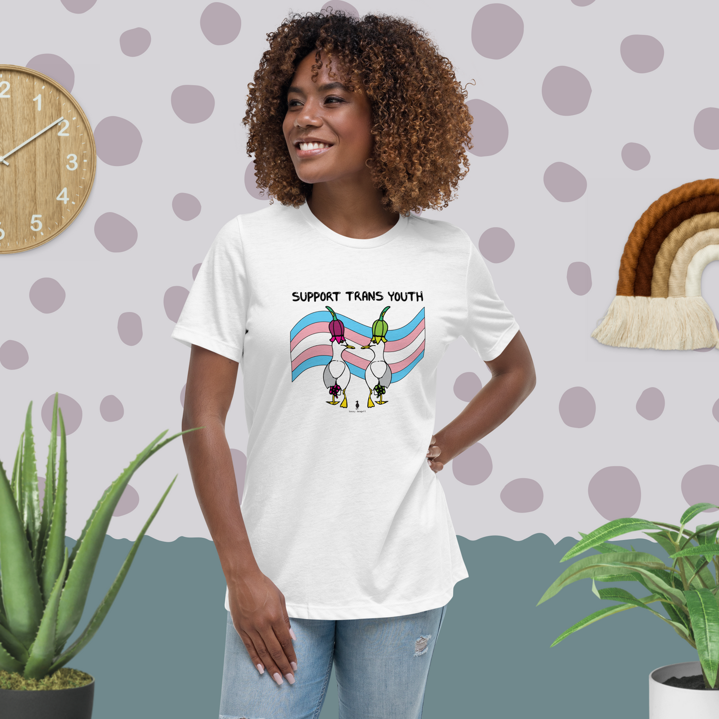 Woman standing facing forwards and looking to the left, wearing a fitted white Sassy Seagull Support Trans Youth LGBTQ+ Pride T-Shirt and pale stonewash blue jeans. She is standing in a pastel coloured boho style room with plants either side and spotty wallpaper. The t-shirt is white with short sleeves. The graphic shows two cartoon style seagulls wearing colourful hats walking away in front of a trans pride flag, underneath the wording Support Trans Youth. It is a soft playful design for everyday wear.